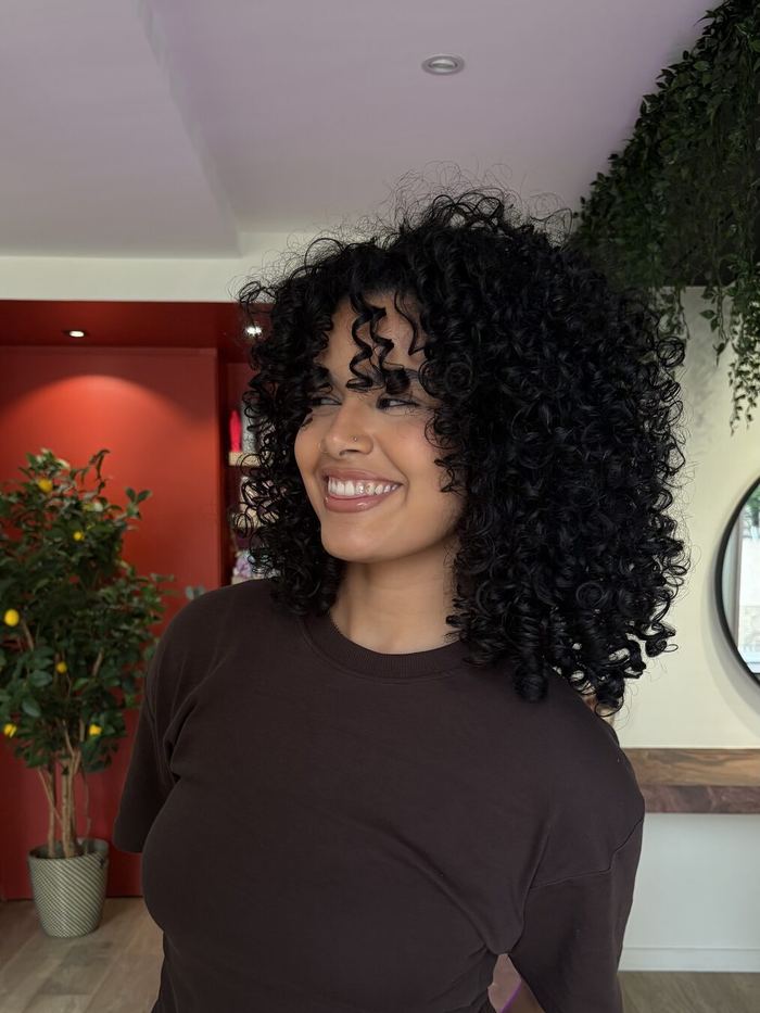 Cliente souriante, carré bouclé noir et frange bouclée, debout dans le salon.
